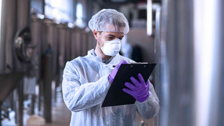 Laboratoriummedewerker in beschermende kleding bekijkt een clipboard terwijl hij langs industriële apparatuur loopt.