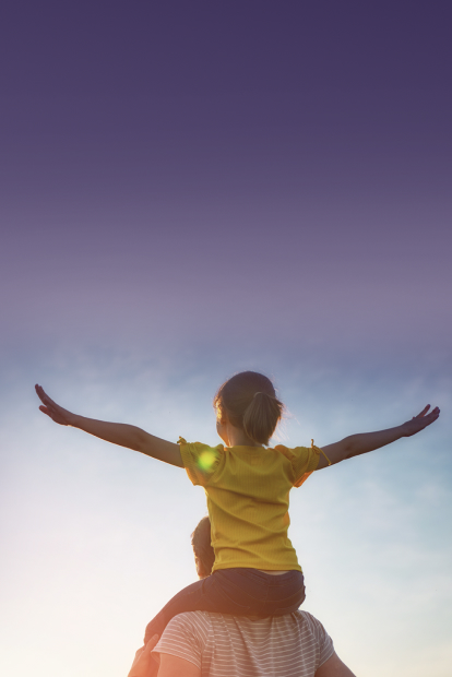 A child sitting on their father’s shoulders looking towards the sun