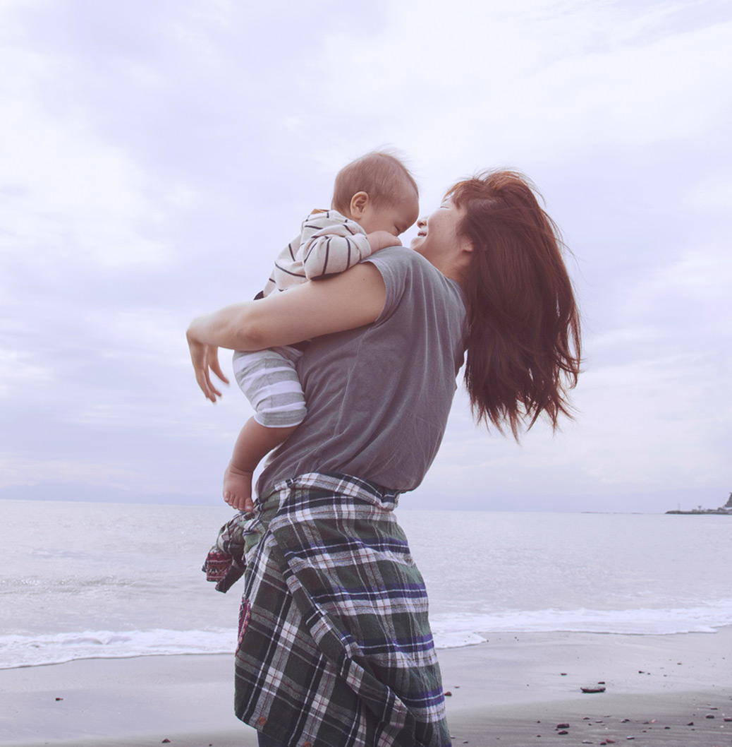 Moeder tilt haar baby op en lacht op het strand, met de zee en een bewolkte hemel op de achtergrond.